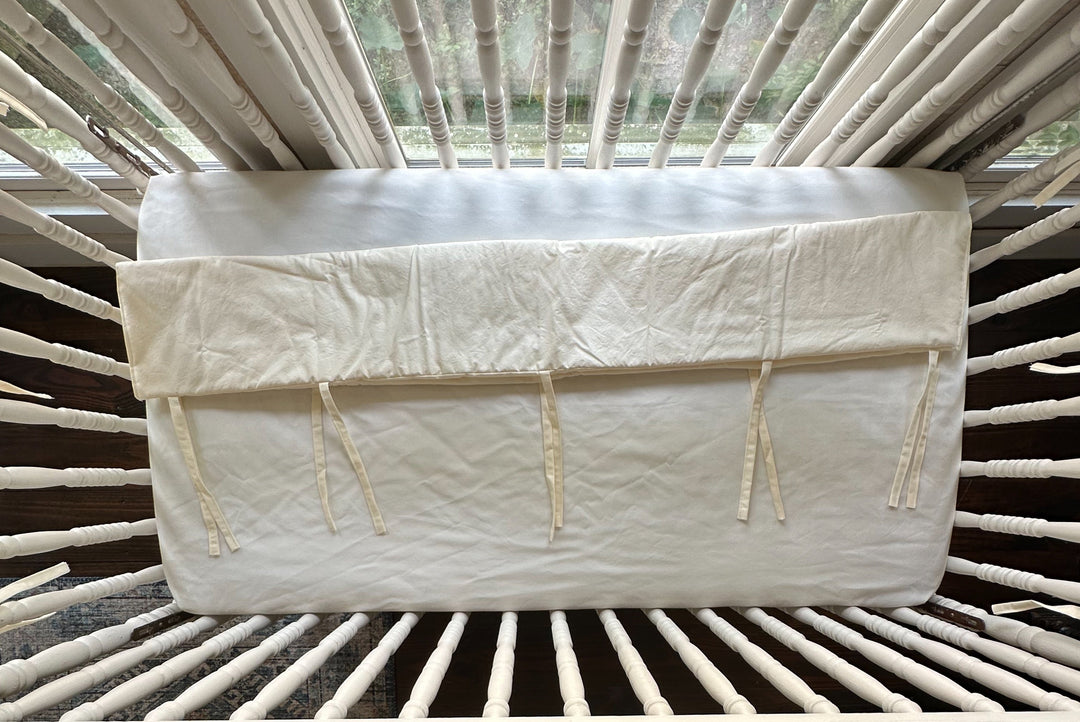 Folded ivory crib rail cover laid across the crib mattress showing its fabric ties