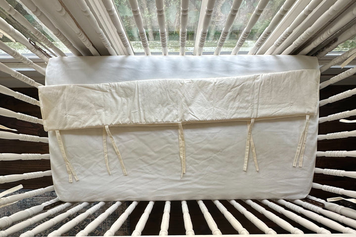 Folded ivory crib rail cover laid across the crib mattress showing its fabric ties