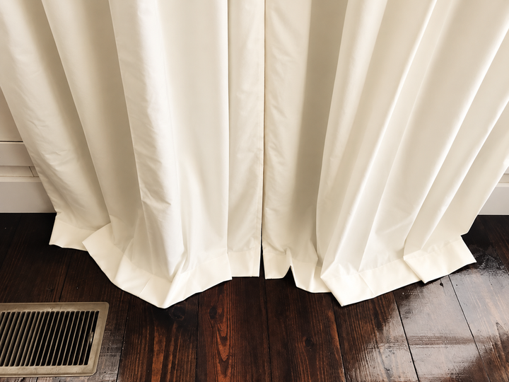Hem detail of ivory blackout curtains breaking against a dark stained wood floor beside a floor vent
