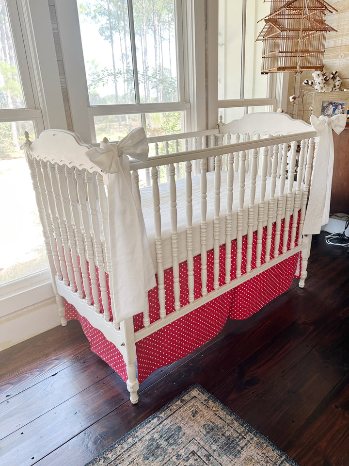 Angled view of the crib with a white sash on the near post and the cherry red polka dot tailored skirt running below the spindles