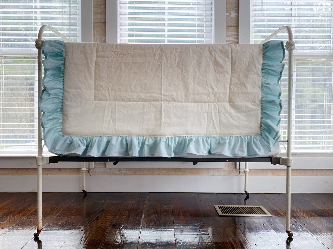 Ivory ruffled baby quilt with a gathered mist blue ruffle draped over an antique iron crib rail