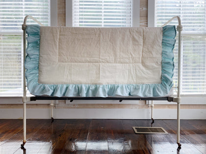 Ivory ruffled baby quilt with a gathered mist blue ruffle draped over an antique iron crib rail