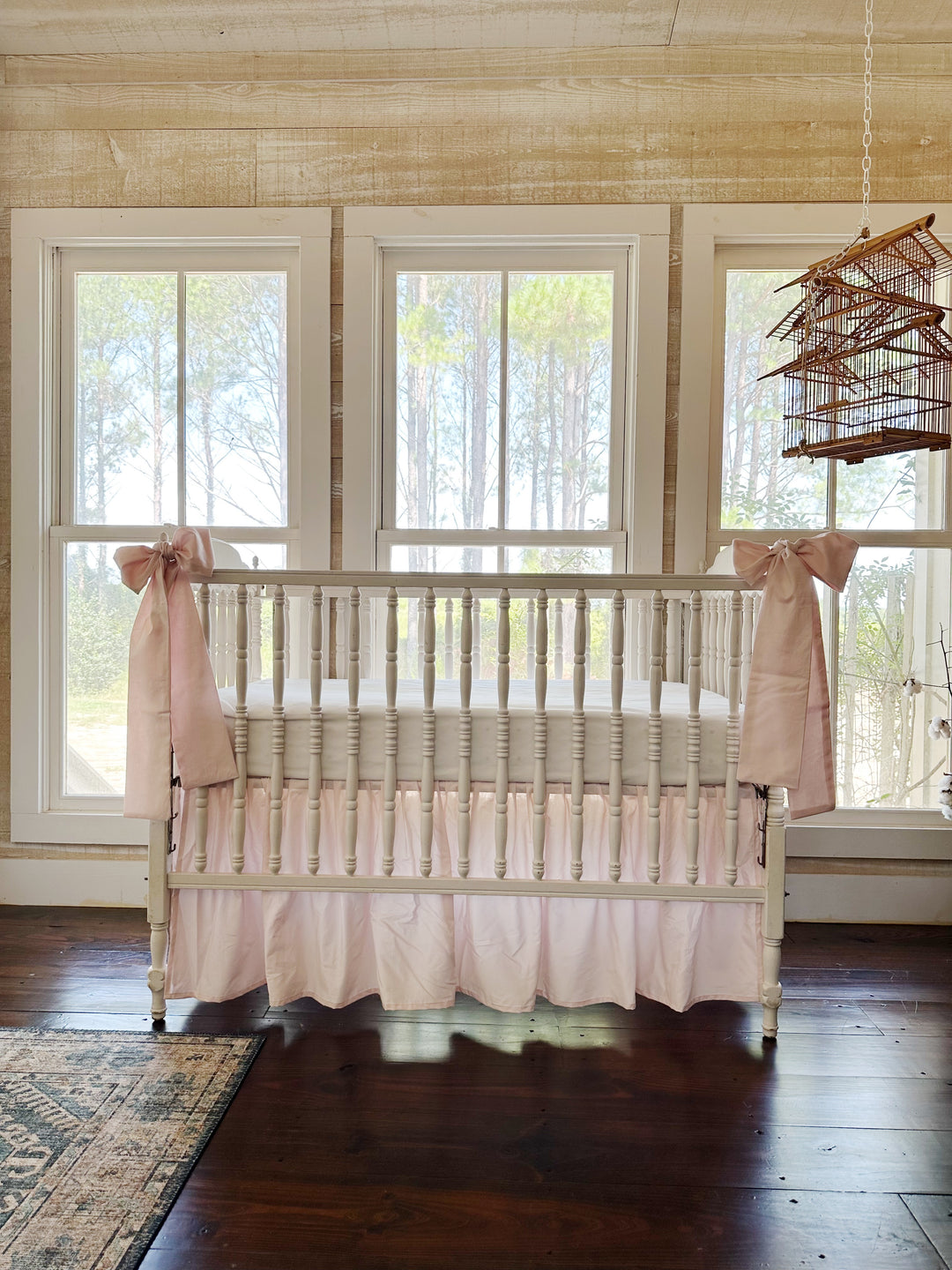 Baby pink gathered farmhouse crib skirt with two large pink bows tied to the front posts of a white spindle crib
