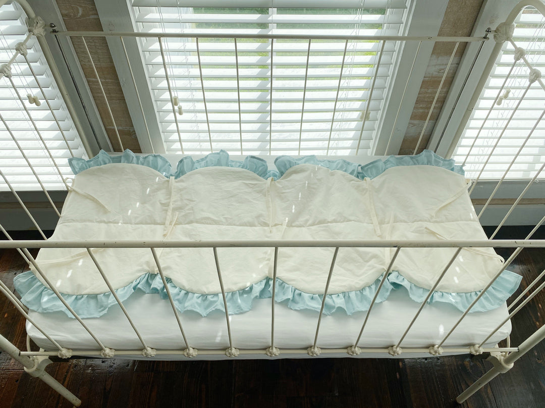 Overhead view of the ivory scalloped crib rail cover showing its full length, mist ruffle and ivory cotton ties