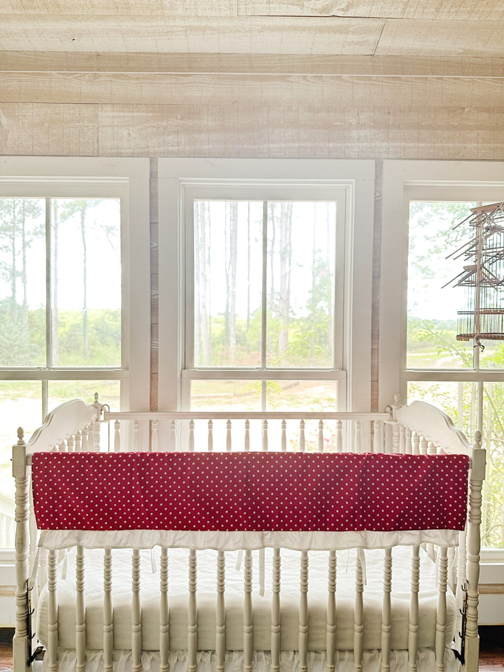 The red polka dot rail cover draped along the top rail of a white Jenny Lind style crib