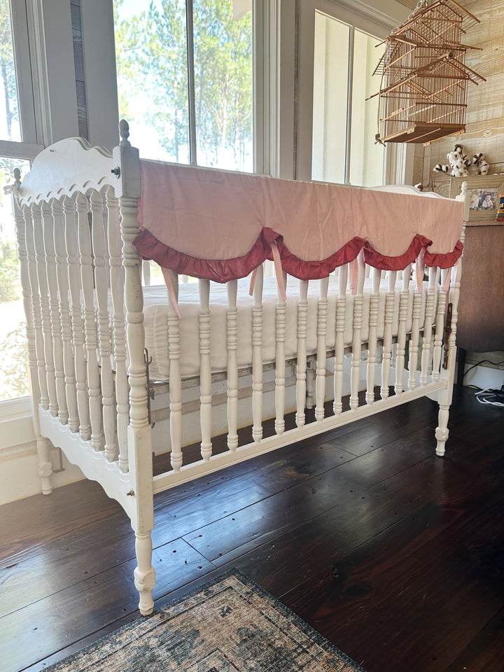 Angled view of the Sisters Pink and Blush scalloped rail cover on a white crib beside an antique birdcage