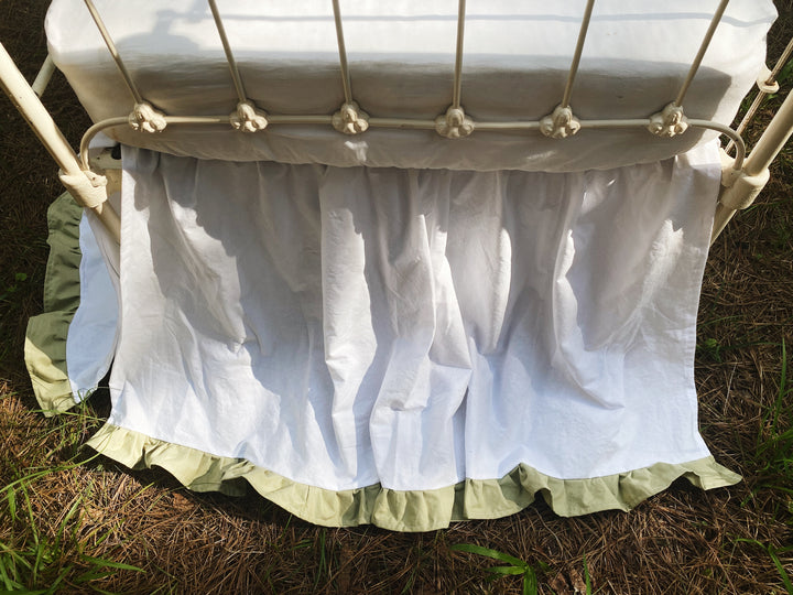 White and sage ruffled crib skirt on antique iron crib in sunlit pine forest, full three-quarter view, handmade nursery bedding