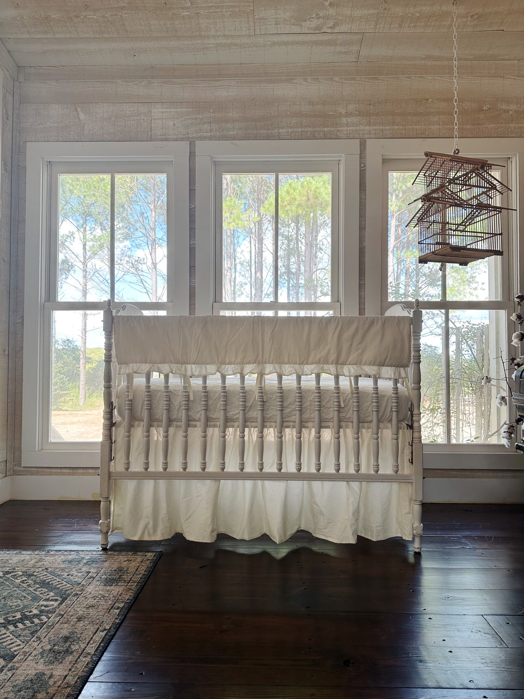 Nursery with whitewashed plank walls and a hanging birdcage, white spindle crib dressed in an ivory ruffled rail cover and gathered ivory skirt
