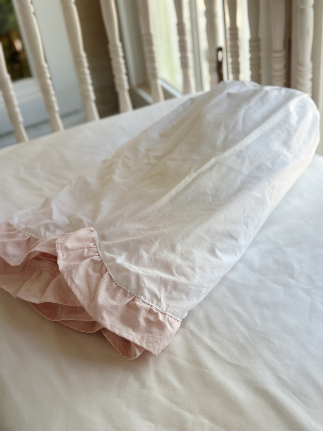 Folded white cotton crib skirt resting on a crib mattress with the baby pink ruffle showing at the fold