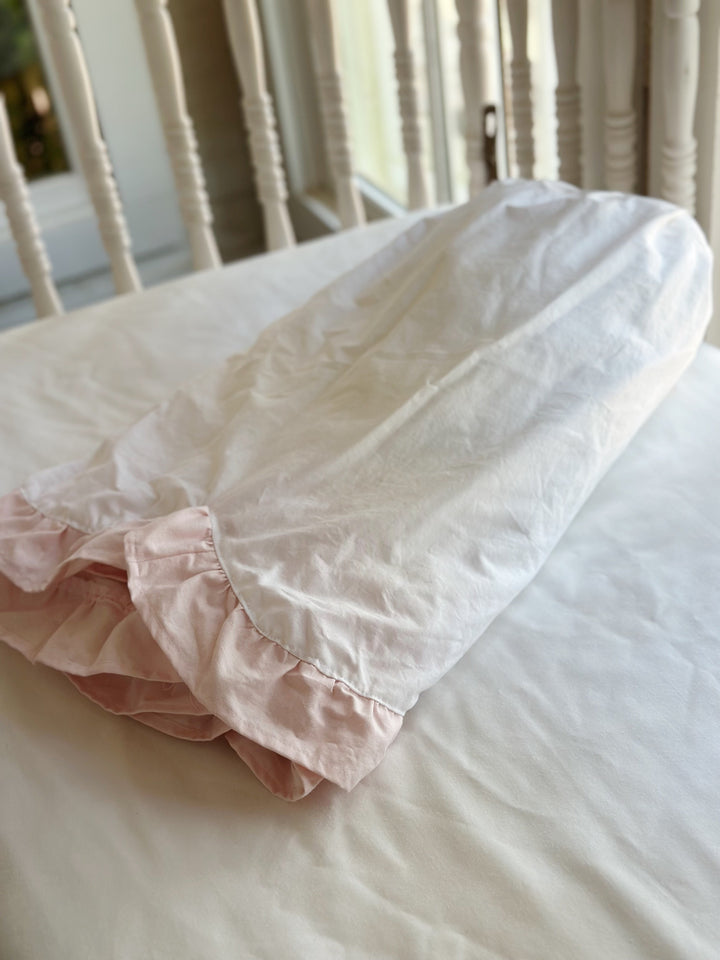 Folded white cotton crib skirt resting on a crib mattress with the baby pink ruffle showing at the fold