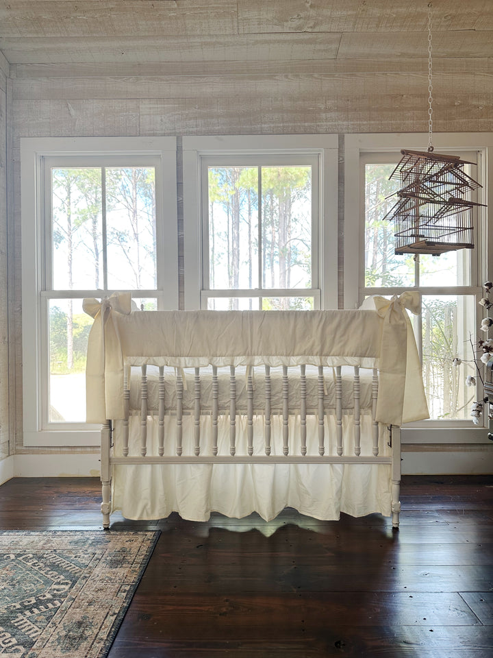 Nursery with whitewashed plank walls and a hanging birdcage, white spindle crib dressed in ivory ruffled rail cover, gathered skirt and corner bows