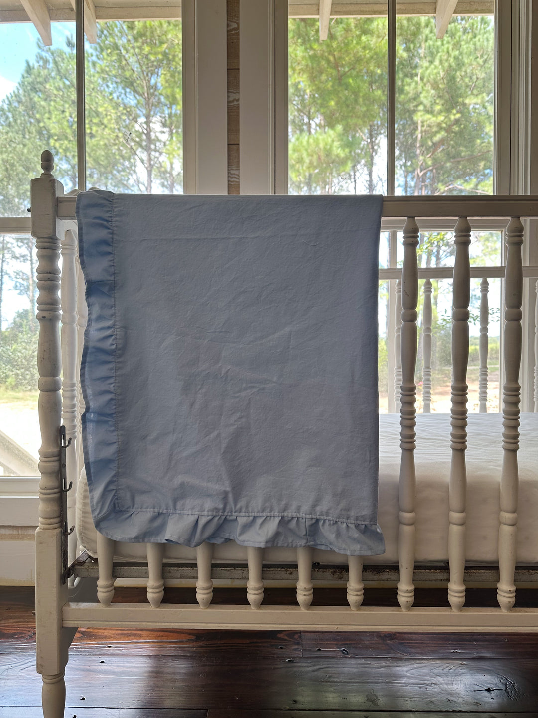 Baby blue ruffled crib blanket hung over the side rail of a white spindle crib in a sunlit nursery - High Cotton Textile