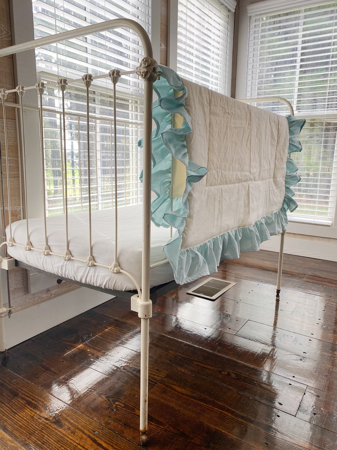 Ivory ruffled crib quilt shown on an iron crib beside a bare white mattress