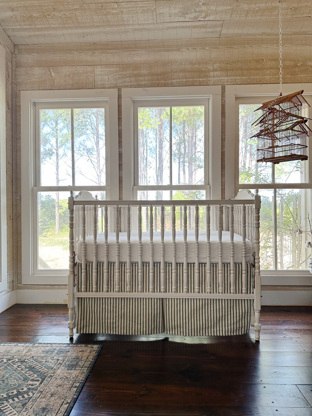 The crib and its green ticking skirt seen across the room, birdcage hanging at the right