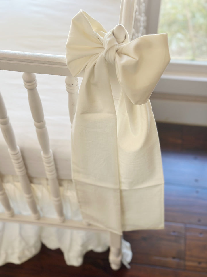 Porcelain crib bow knotted around the finial of the crib's corner post, long tail hanging past the ruffled skirt