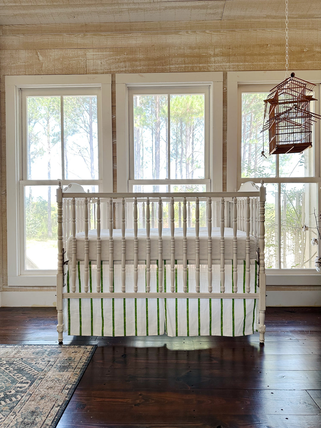 The striped crib skirt seen across a pine-paneled room with three windows and a hanging birdcage