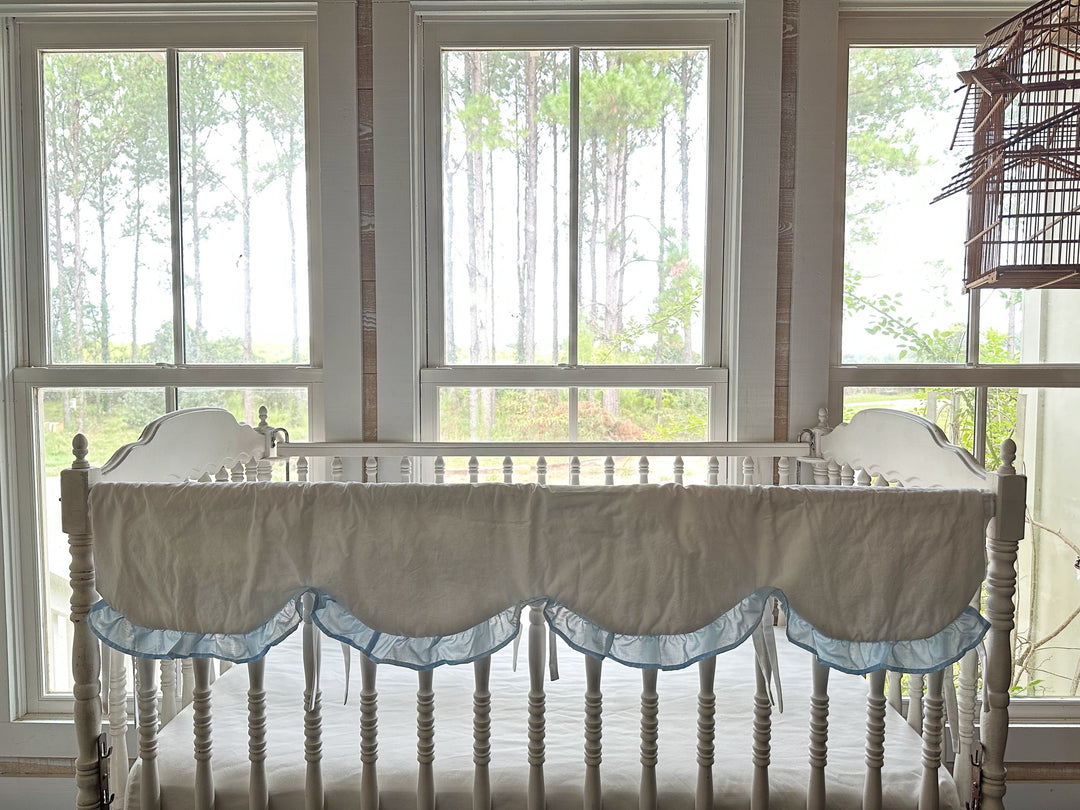 White scalloped crib rail cover with heirloom blue ruffle tied along the front rail of a spindle crib