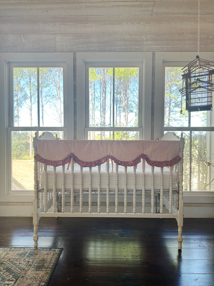 Wide nursery view of the Sisters Pink and Blush rail cover on a white crib in front of three tall windows
