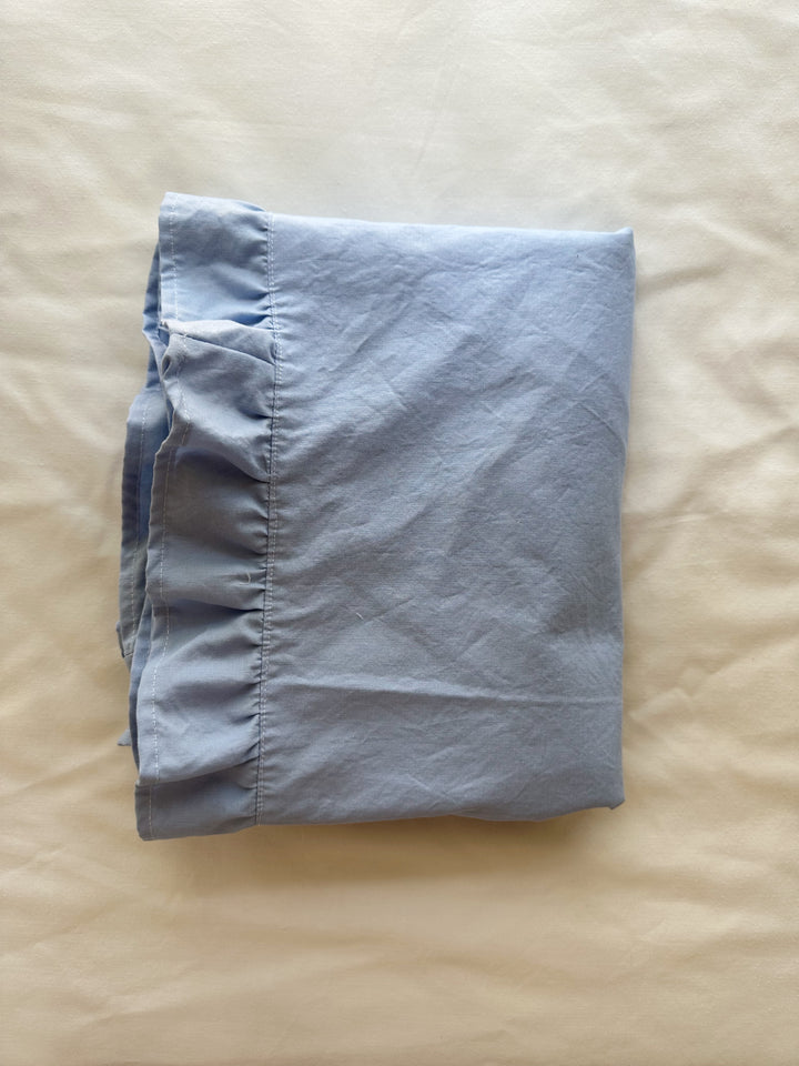 Baby blue ruffled crib blanket folded on cream linen, showing the ruffle border and hem - High Cotton Textile