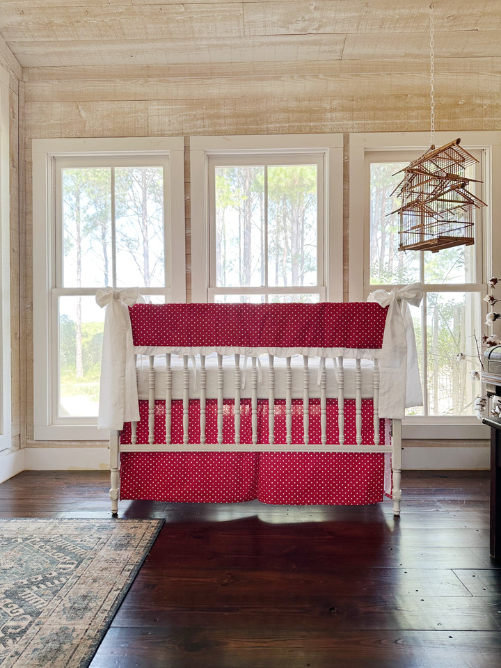 The red polka dot crib set in a pine-paneled nursery with three tall windows and a hanging birdcage