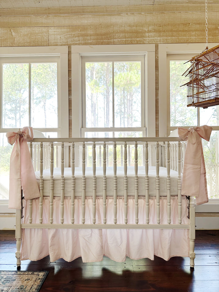 The baby pink crib skirt and bows in a pine-paneled nursery with three tall windows and a hanging birdcage