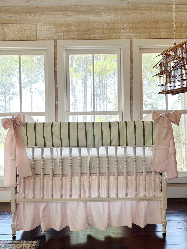 Green and white striped tailored crib rail cover with a baby pink farmhouse skirt and two pink bows on a white spindle crib