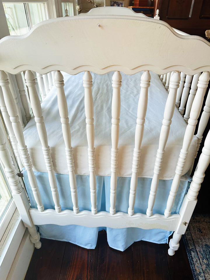 Foot of the crib with its scalloped end board, showing the heirloom blue skirt's center pleat and split corners at the legs - High Cotton Textile