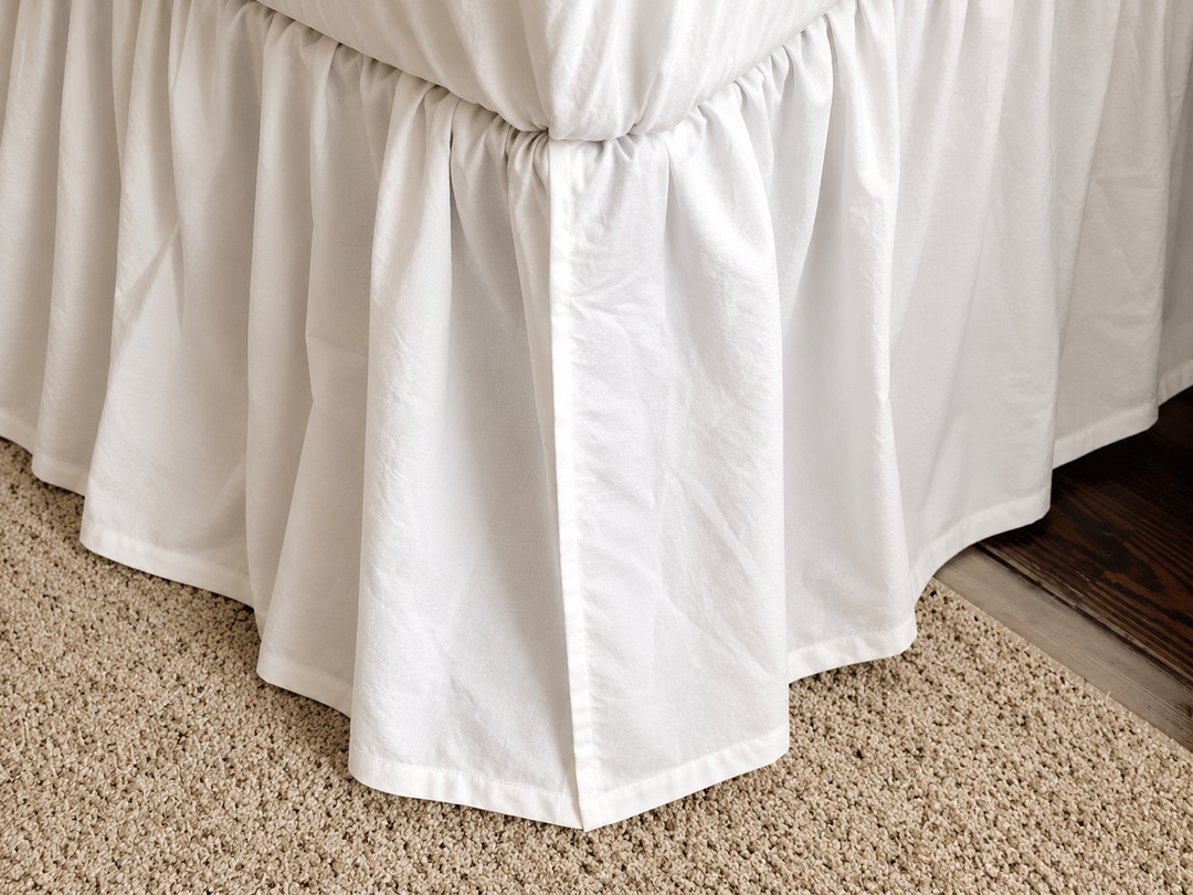 Split corner at the foot of the white farmhouse bed skirt, where the two gathered panels overlap around a footboard post