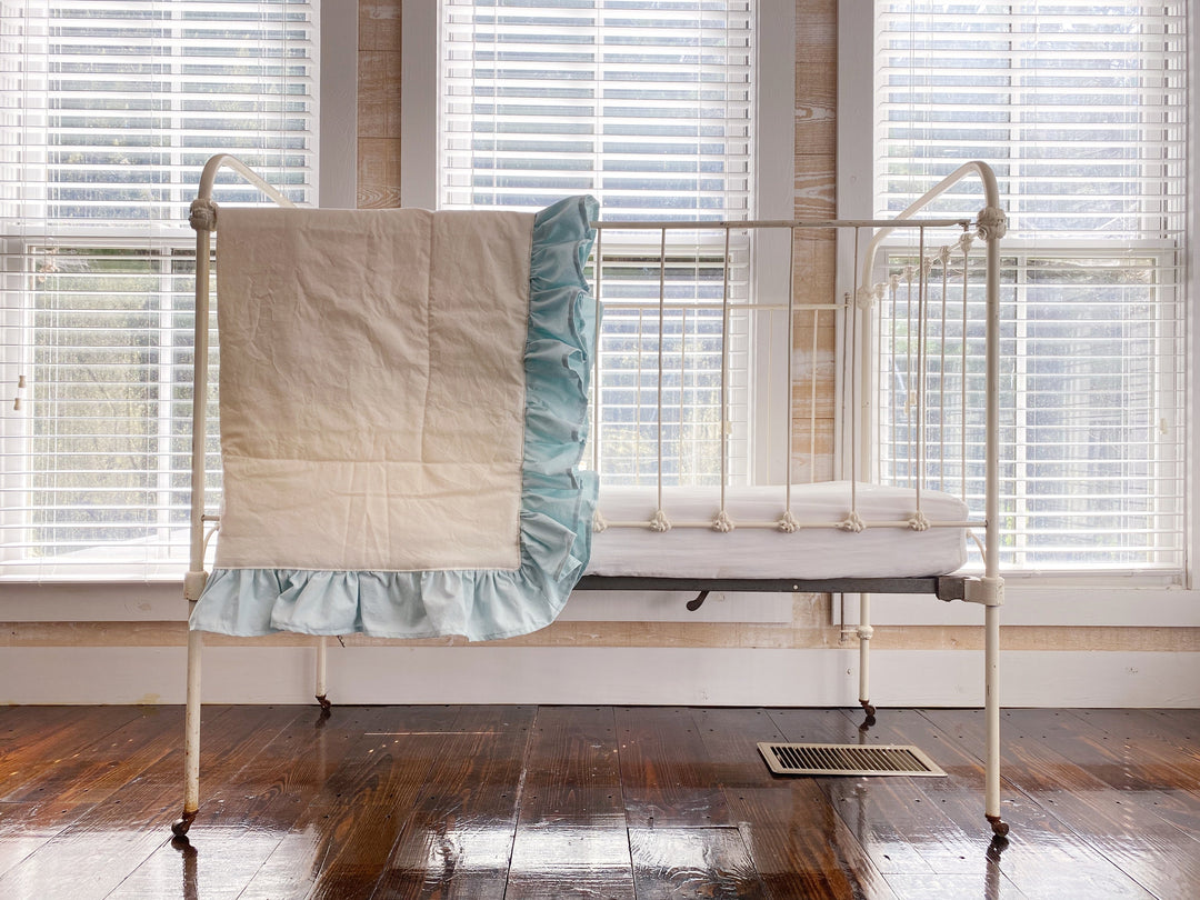 Wide view of an iron crib with the ivory and mist ruffled quilt draped over one end