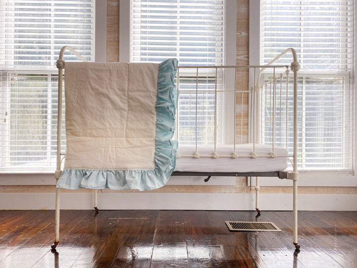 Wide view of an iron crib with the ivory and mist ruffled quilt draped over one end