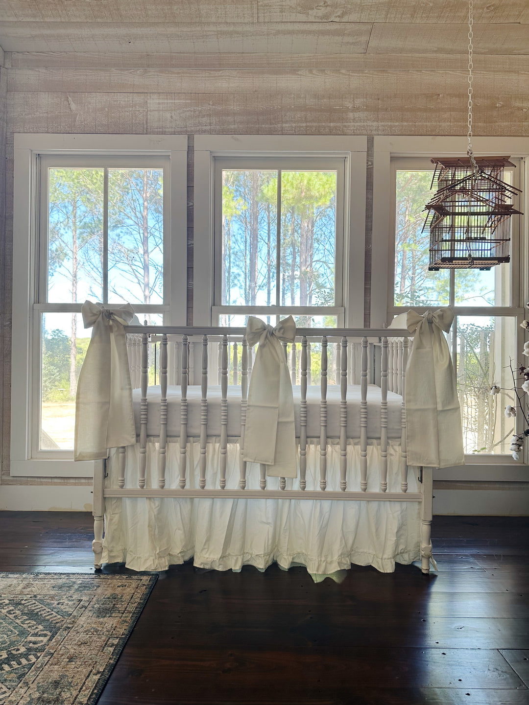 Nursery with shiplap walls and a hanging birdcage, white spindle crib dressed in a porcelain ruffled skirt with bows at both corners and center