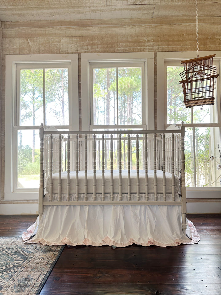 Spindle crib dressed in the white and baby pink scalloped skirt in a nursery with pine views