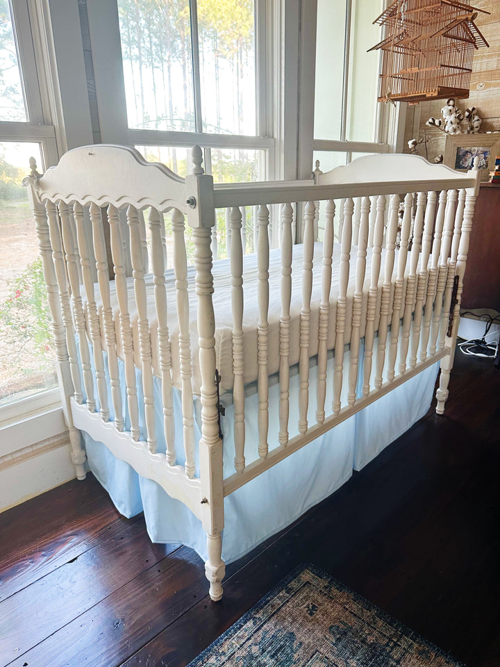 Angled view of the heirloom blue crib skirt wrapping the side and foot of a white spool-spindle crib beneath a hanging birdcage - High Cotton Textile