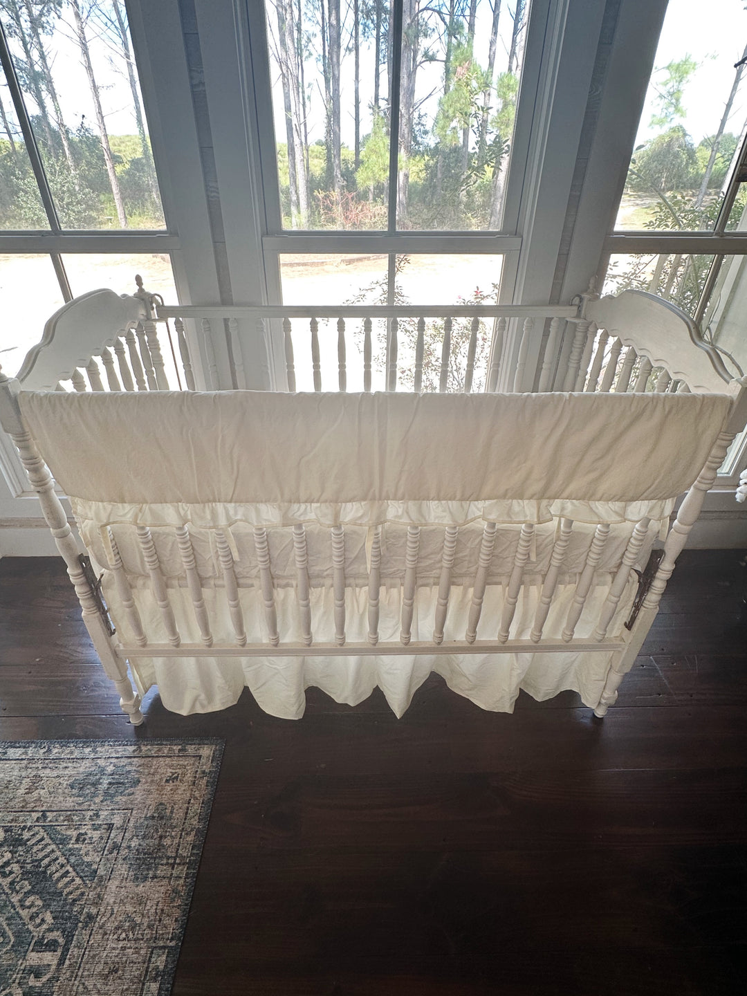 Backlit front view of the ivory ruffled rail cover across the top rail, farmhouse skirt gathered to dark wood floors