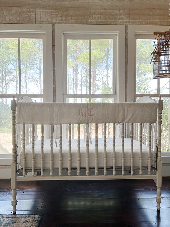 White simple crib rail cover with a pink monogram tied along the front rail of a spindle crib