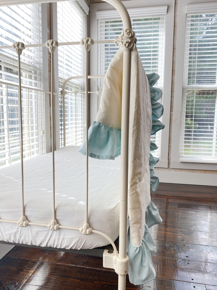 Ivory baby quilt with mist ruffle draped over the corner post of a vintage iron crib