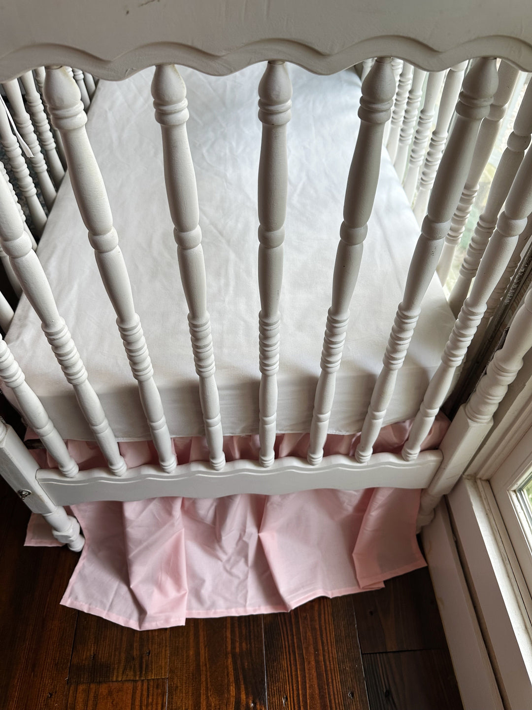 Sisters Pink farmhouse crib skirt with split corners under a white fitted crib sheet on a white spindle crib