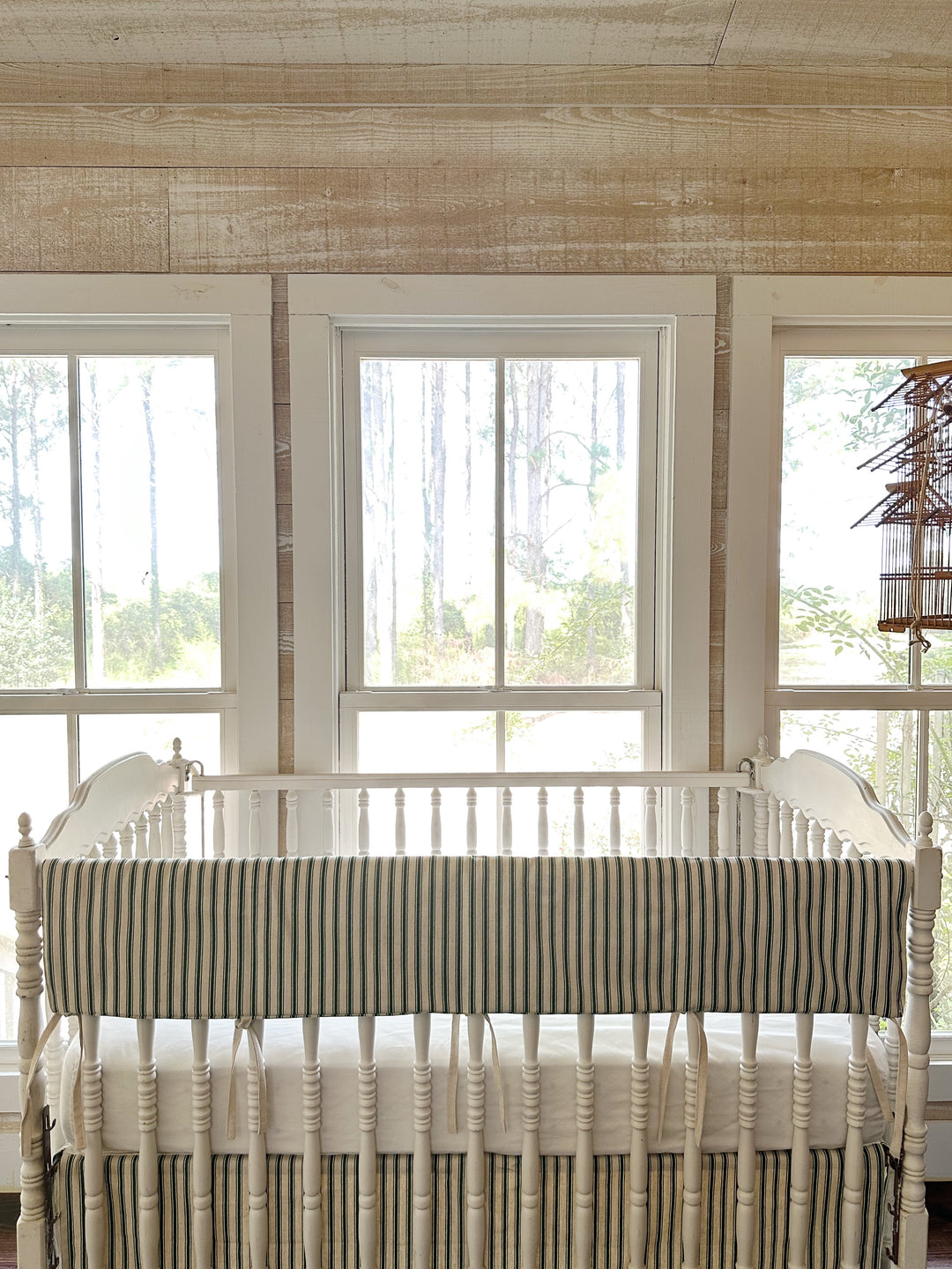 The ticking rail cover on the crib in a pine-paneled room with tall windows and a hanging birdcage