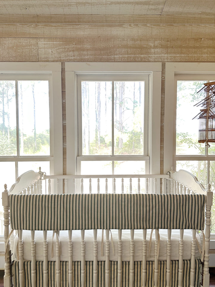 The ticking rail cover on the crib in a pine-paneled room with tall windows and a hanging birdcage