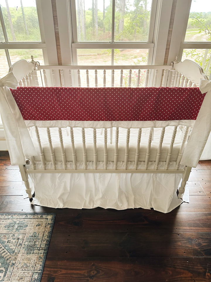 Red and white polka dot ruffled crib rail cover with a white farmhouse skirt and two white bows on a spindle crib
