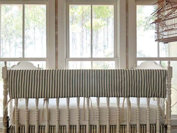 Pine green ticking simple crib rail cover across the long rail of a white spindle crib, natural ties hanging below