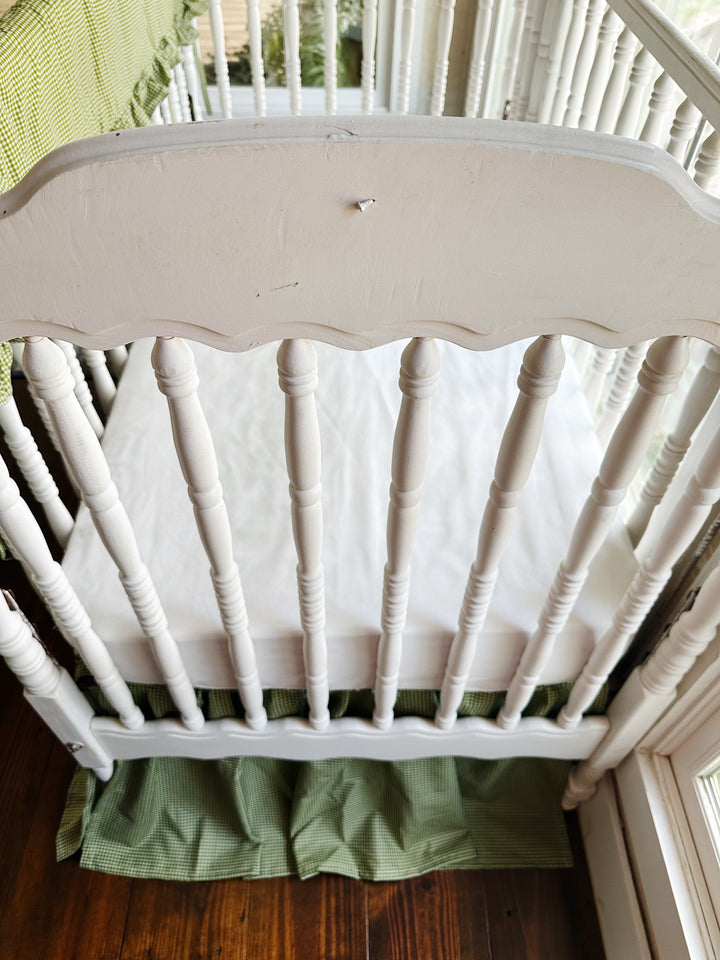 End view of white spindle crib with green gingham crib skirt and white crib sheet