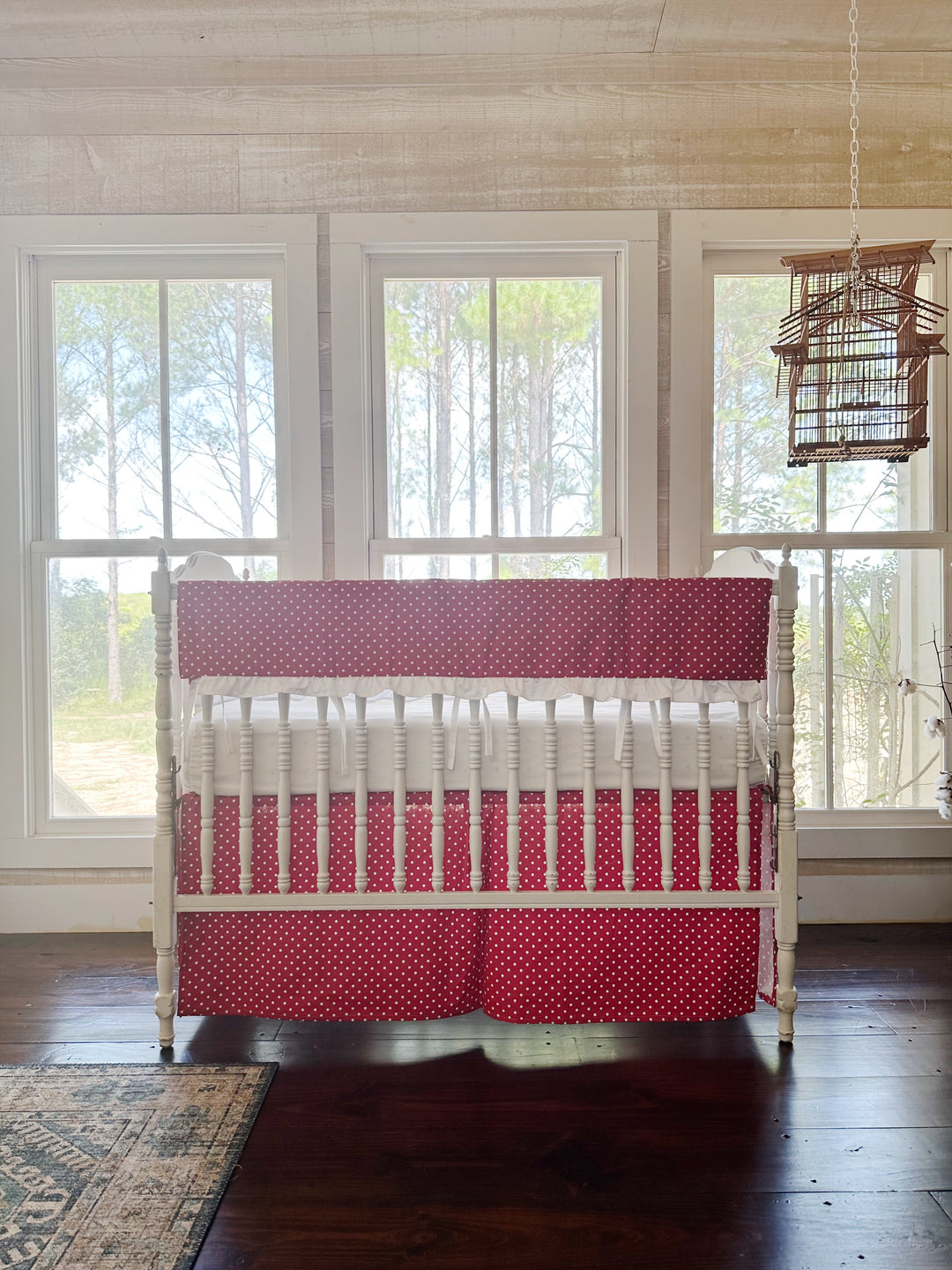 The red polka dot crib set in a pine-paneled nursery with three tall windows and a hanging birdcage