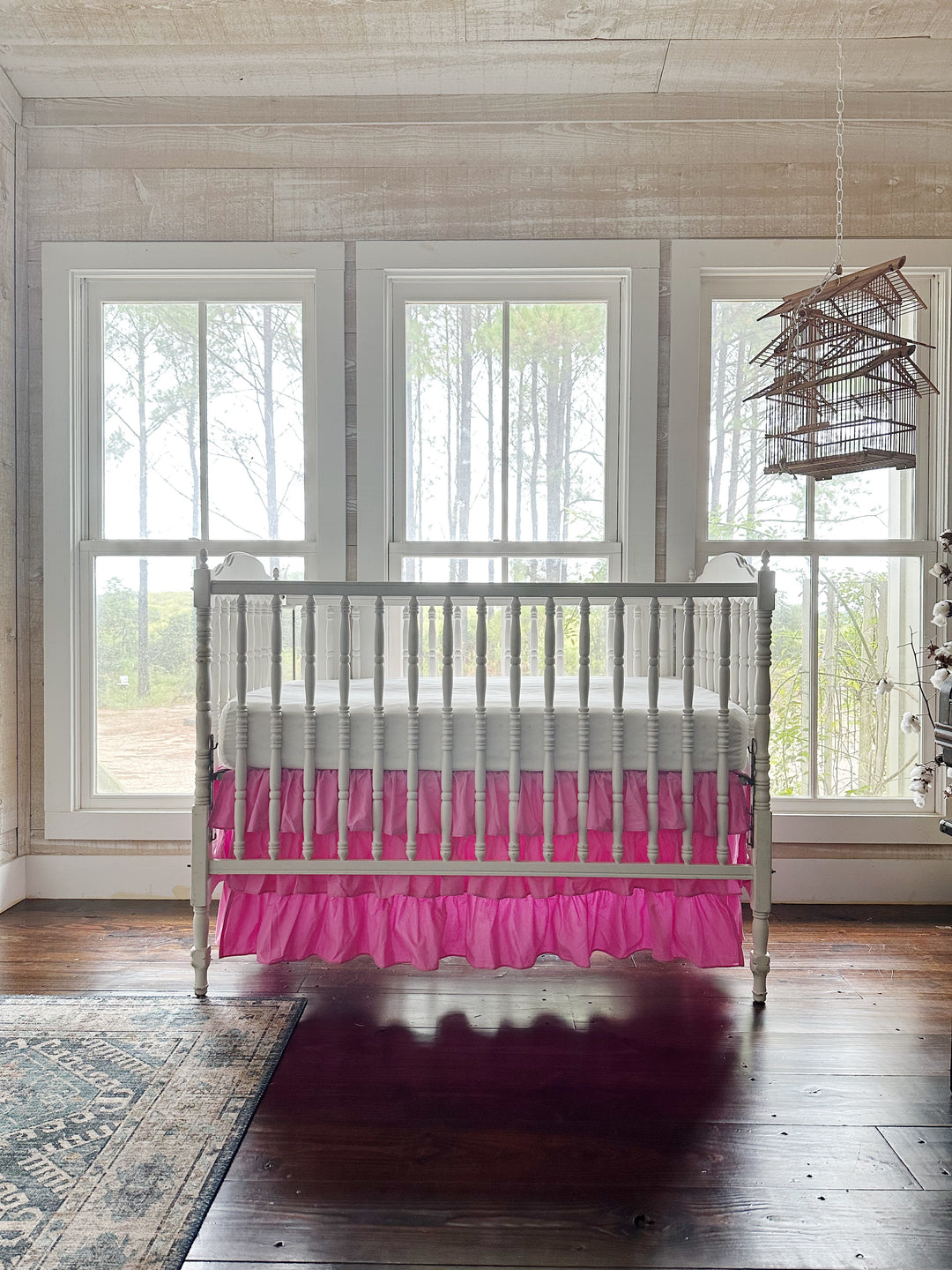 Nursery with whitewashed plank walls and a hanging birdcage, white spindle crib dressed in a pink three-tier ruffled crib skirt