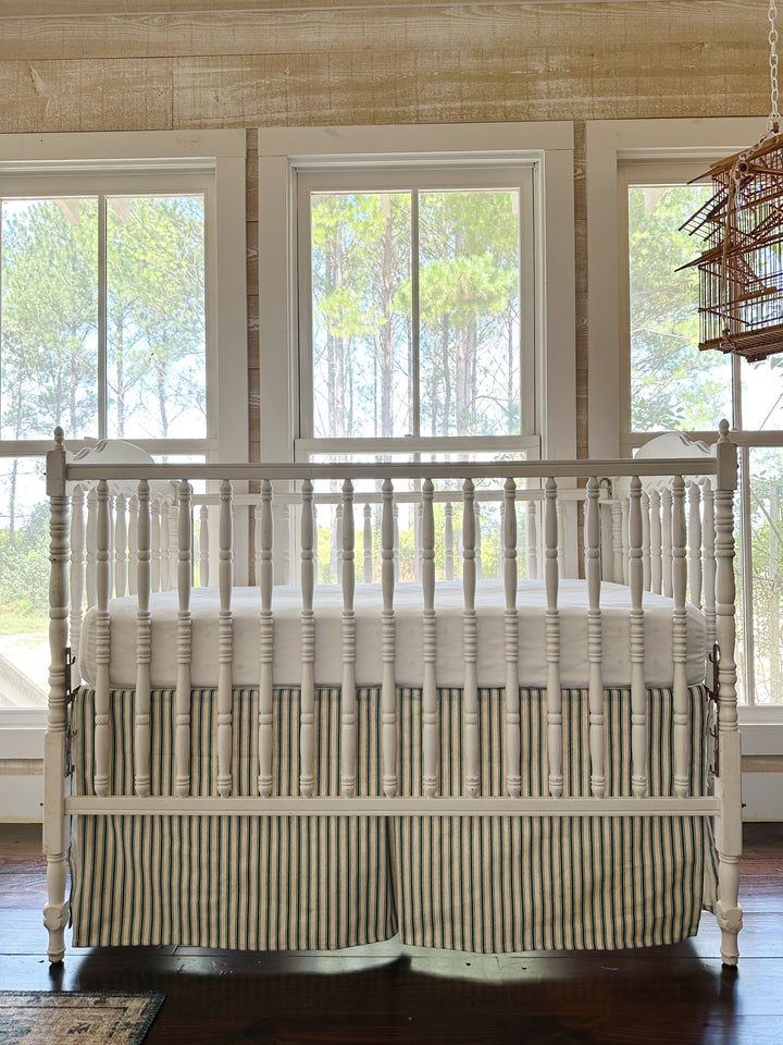 The striped skirt straight on beneath a white crib, pine-paneled wall and three windows behind