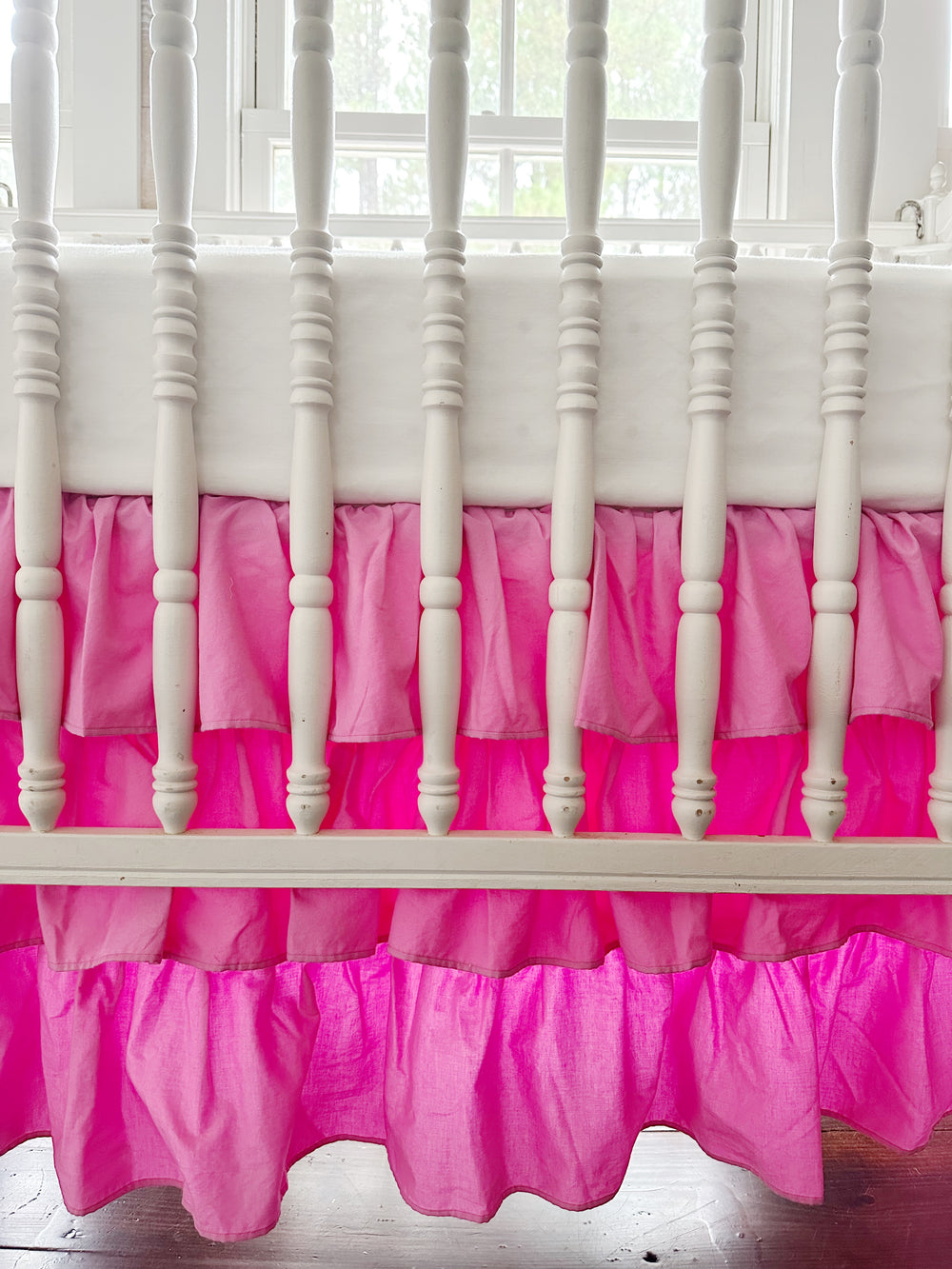Close-up of the three gathered parfait pink tiers seen through the white crib spindles, window light glowing through the lower ruffles
