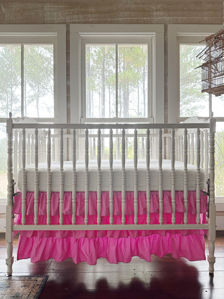 Parfait pink tiered ruffled crib skirt with three layers of ruffles on a white spindle crib, straight-on in front of three tall windows