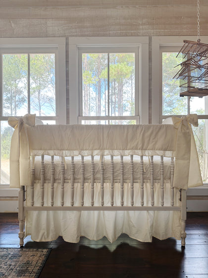 Ivory ruffled crib rail cover, farmhouse crib skirt and two ivory bows on a white spindle crib, straight-on in front of three tall windows