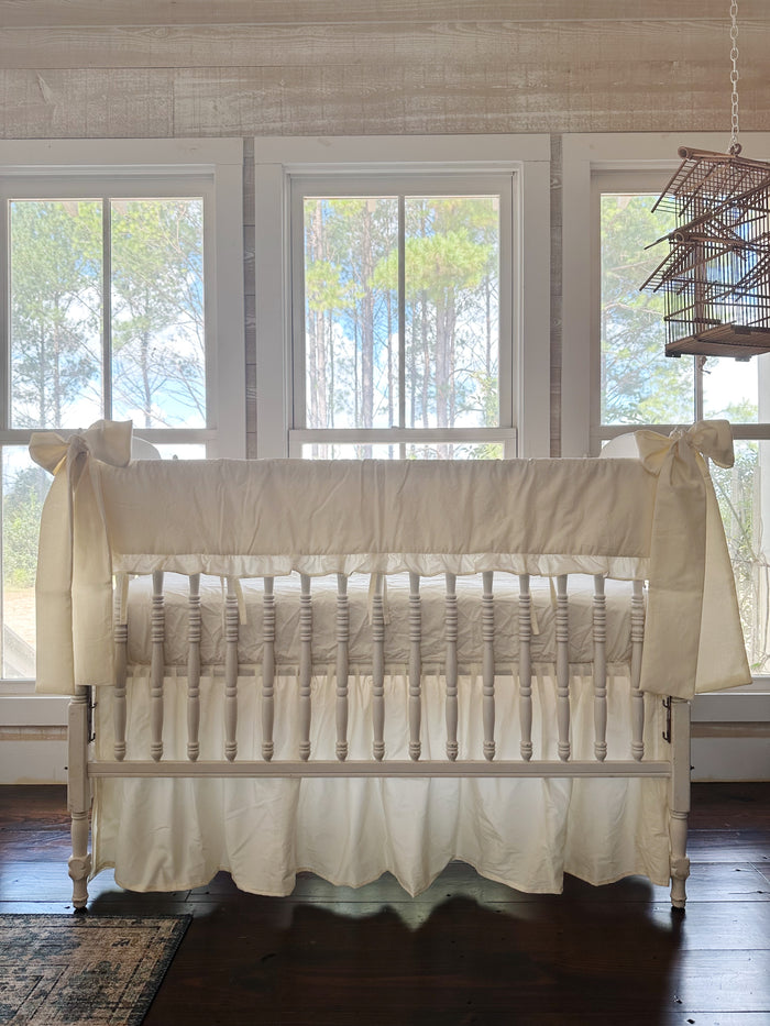 Ivory ruffled crib rail cover, farmhouse crib skirt and two ivory bows on a white spindle crib, straight-on in front of three tall windows