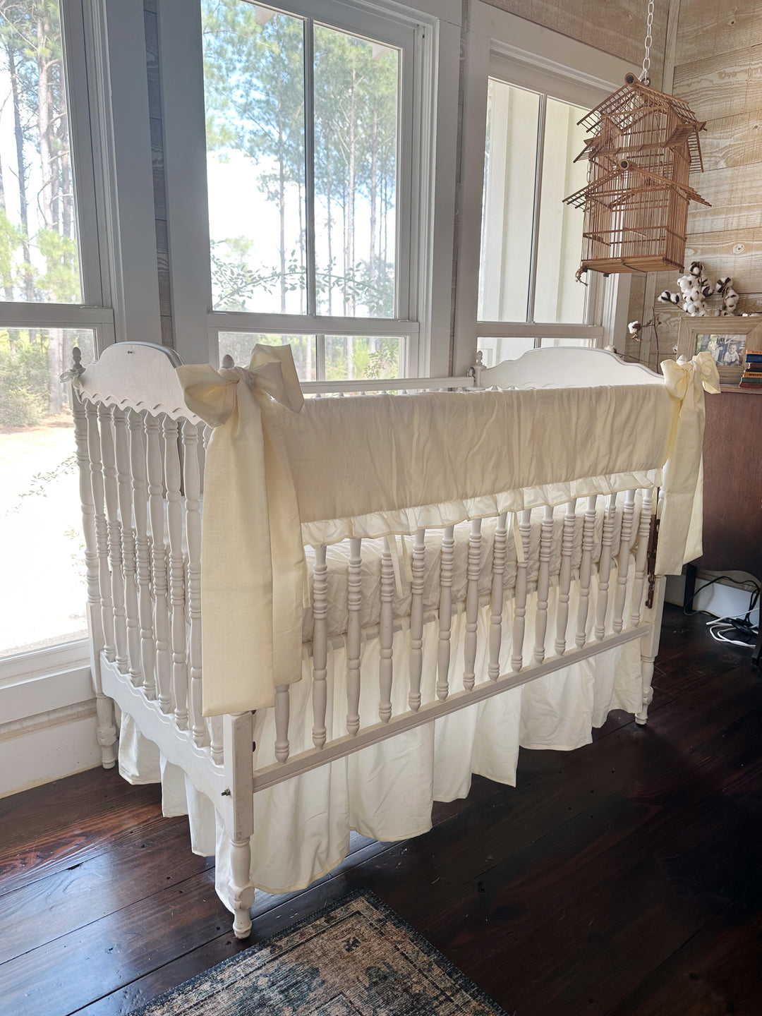 Three-quarter view of the ivory crib bedding set by a bank of windows, bows tied at both front corner posts